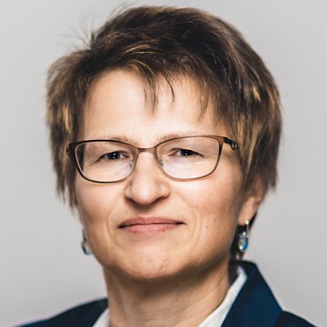 Beate Ulbrich hat kurzes dunkelblondes Haar, sie trägt eine Brille, Ohrringe, eine weiße Bluse und ein dunkelblaues Jackett.