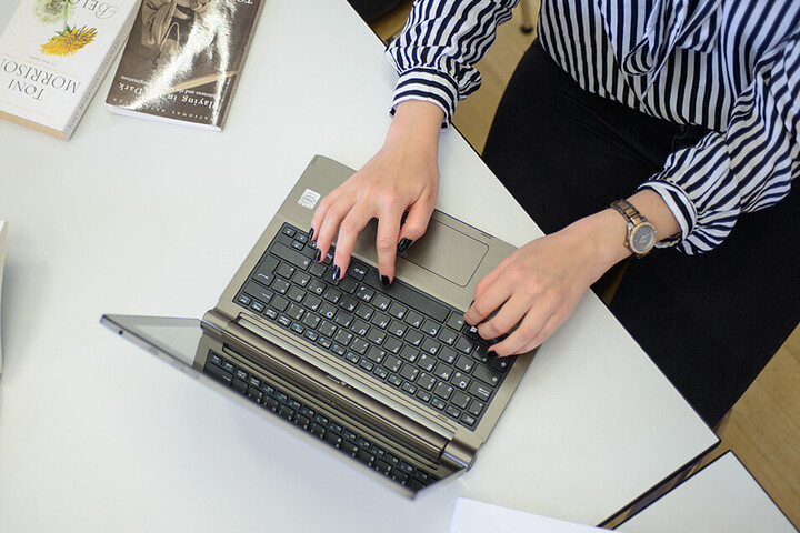 Eine Frau schreibt an einem Laptop. Sie trägt eine schwarz-weiß gemusterte Bluse, eine schwarze Hose und am rechten Handgelenk eine Uhr. Ihre Fingernägel sind schwarz lackiert. Neben dem Laptop liegen ein aufgeschlagenes Buch, auf dem ein Stift und eine Brille abgelegt sind, weitere Bücher und ein schwarzes Schlüsselband mit weißem Aufdruck "Universität Mannheim" und dem Logo.