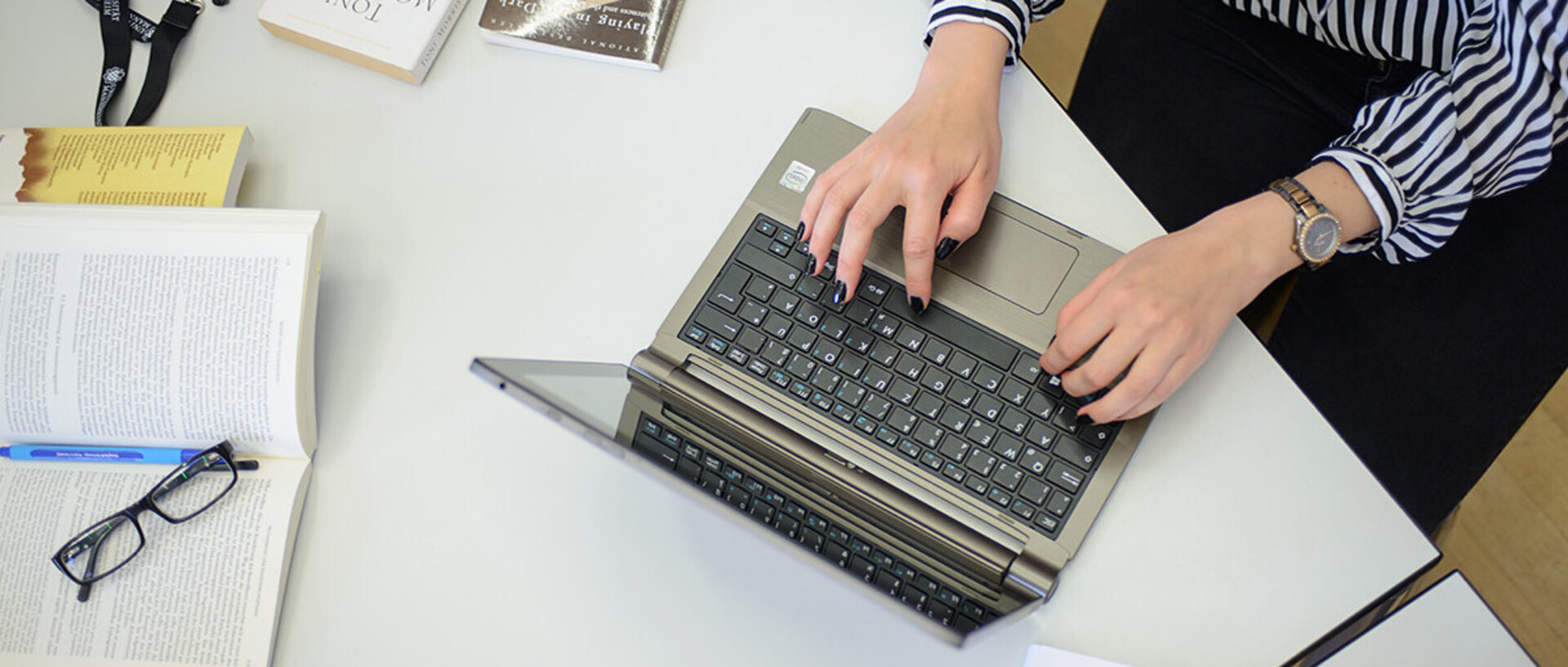 Eine Frau schreibt an einem Laptop. Sie trägt eine schwarz-weiß gemusterte Bluse, eine schwarze Hose und am rechten Handgelenk eine Uhr. Ihre Fingernägel sind schwarz lackiert. Neben dem Laptop liegen ein aufgeschlagenes Buch, auf dem ein Stift und eine Brille abgelegt sind, weitere Bücher und ein schwarzes Schlüsselband mit weißem Aufdruck "Universität Mannheim" und dem Logo.