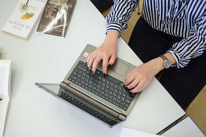 Eine Frau schreibt an einem Laptop. Sie trägt eine schwarz-weiß gemusterte Bluse, eine schwarze Hose und am rechten Handgelenk eine Uhr. Ihre Fingernägel sind schwarz lackiert. Neben dem Laptop liegen ein aufgeschlagenes Buch, auf dem ein Stift und eine Brille abgelegt sind, weitere Bücher und ein schwarzes Schlüsselband mit weißem Aufdruck "Universität Mannheim" und dem Logo.