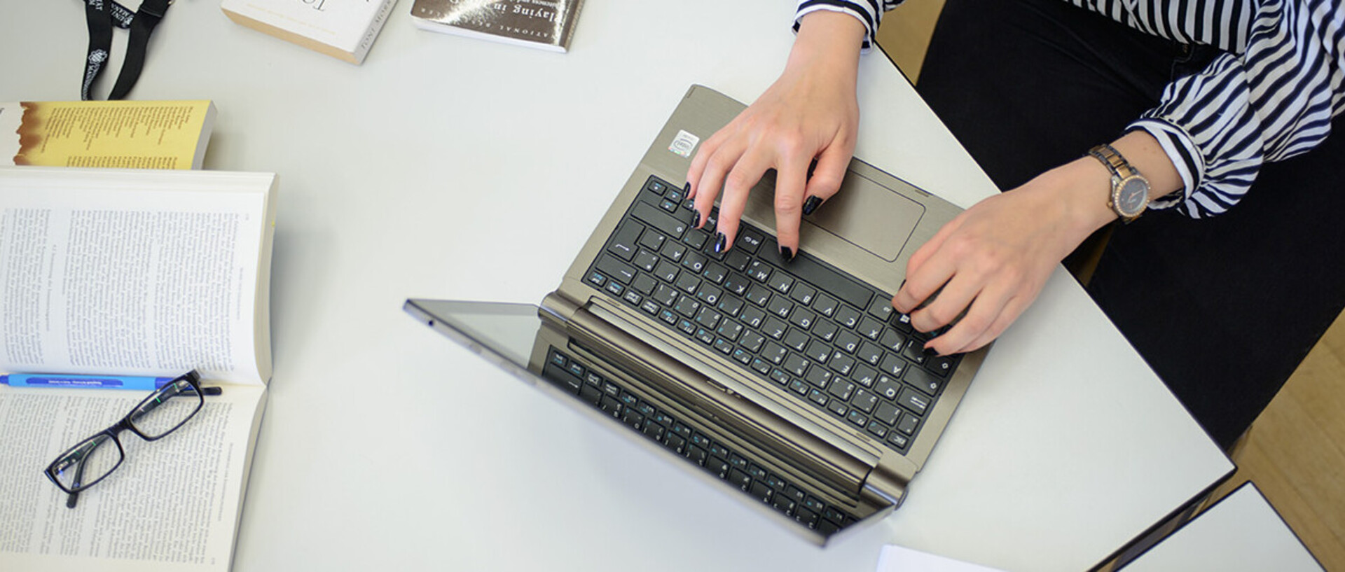 Eine Frau schreibt an einem Laptop. Sie trägt eine schwarz-weiß gemusterte Bluse, eine schwarze Hose und am rechten Handgelenk eine Uhr. Ihre Fingernägel sind schwarz lackiert. Neben dem Laptop liegen ein aufgeschlagenes Buch, auf dem ein Stift und eine Brille abgelegt sind, weitere Bücher und ein schwarzes Schlüsselband mit weißem Aufdruck "Universität Mannheim" und dem Logo.
