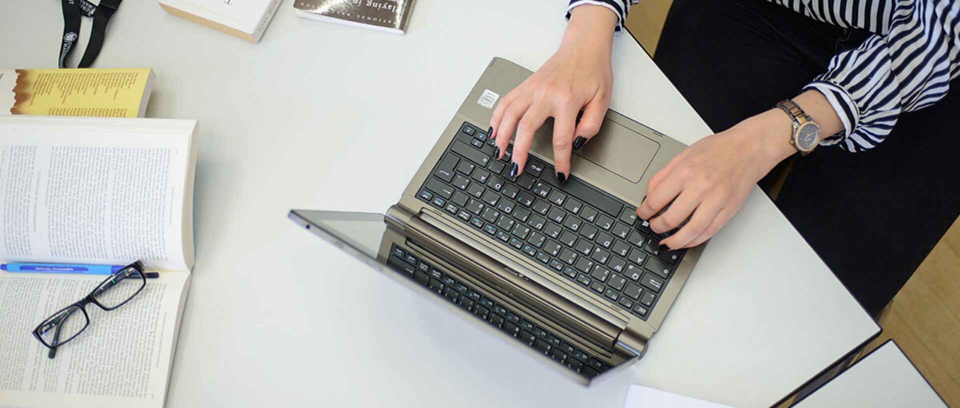 Eine Frau schreibt an einem Laptop. Sie trägt eine schwarz-weiß gemusterte Bluse, eine schwarze Hose und am rechten Handgelenk eine Uhr. Ihre Fingernägel sind schwarz lackiert. Neben dem Laptop liegen ein aufgeschlagenes Buch, auf dem ein Stift und eine Brille abgelegt sind, weitere Bücher und ein schwarzes Schlüsselband mit weißem Aufdruck "Universität Mannheim" und dem Logo.