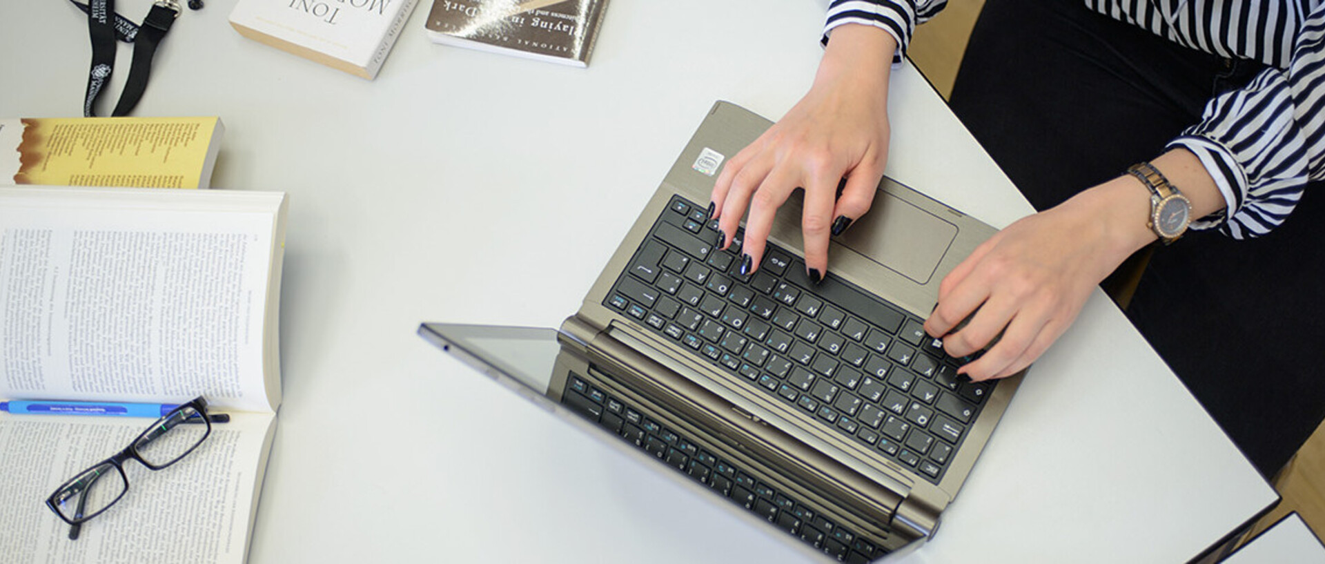 Eine Frau schreibt an einem Laptop. Sie trägt eine schwarz-weiß gemusterte Bluse, eine schwarze Hose und am rechten Handgelenk eine Uhr. Ihre Fingernägel sind schwarz lackiert. Neben dem Laptop liegen ein aufgeschlagenes Buch, auf dem ein Stift und eine Brille abgelegt sind, weitere Bücher und ein schwarzes Schlüsselband mit weißem Aufdruck "Universität Mannheim" und dem Logo.
