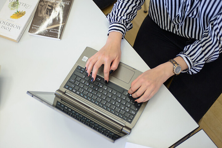 Eine Frau schreibt an einem Laptop. Sie trägt eine schwarz-weiß gemusterte Bluse, eine schwarze Hose und am rechten Handgelenk eine Uhr. Ihre Fingernägel sind schwarz lackiert. Neben dem Laptop liegen ein aufgeschlagenes Buch, auf dem ein Stift und eine Brille abgelegt sind, weitere Bücher und ein schwarzes Schlüsselband mit weißem Aufdruck "Universität Mannheim" und dem Logo.